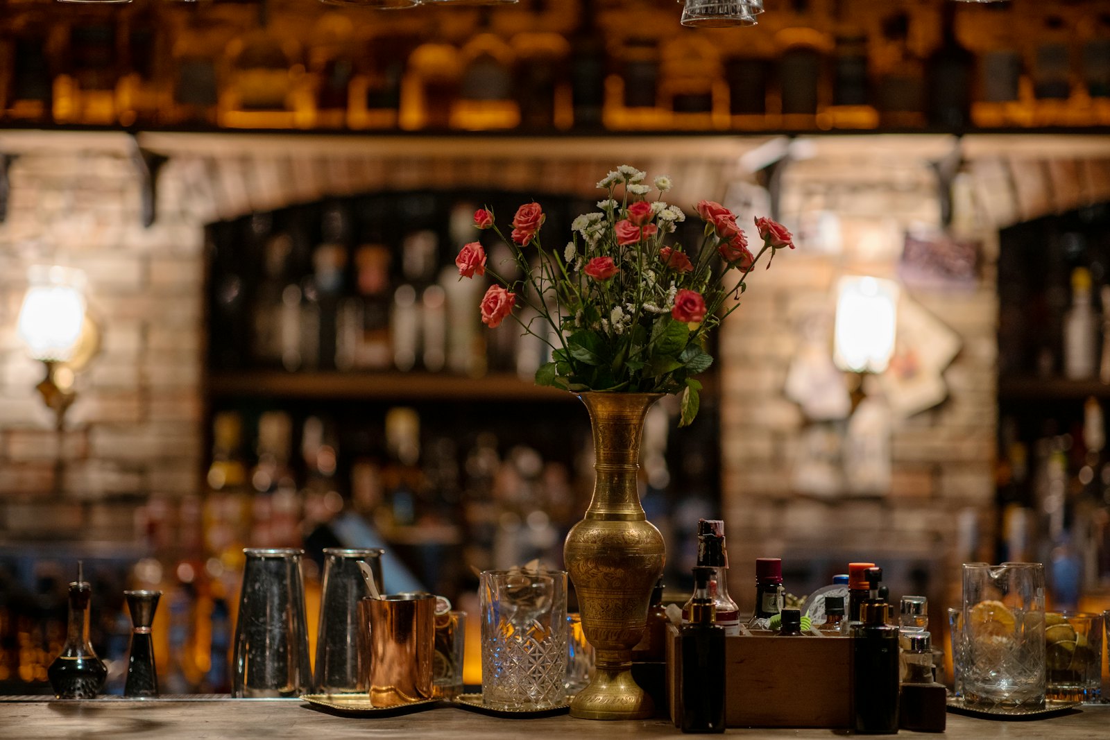 A bar counter set with mixing glasses, bitters and a brass vase of roses, lit warm at night